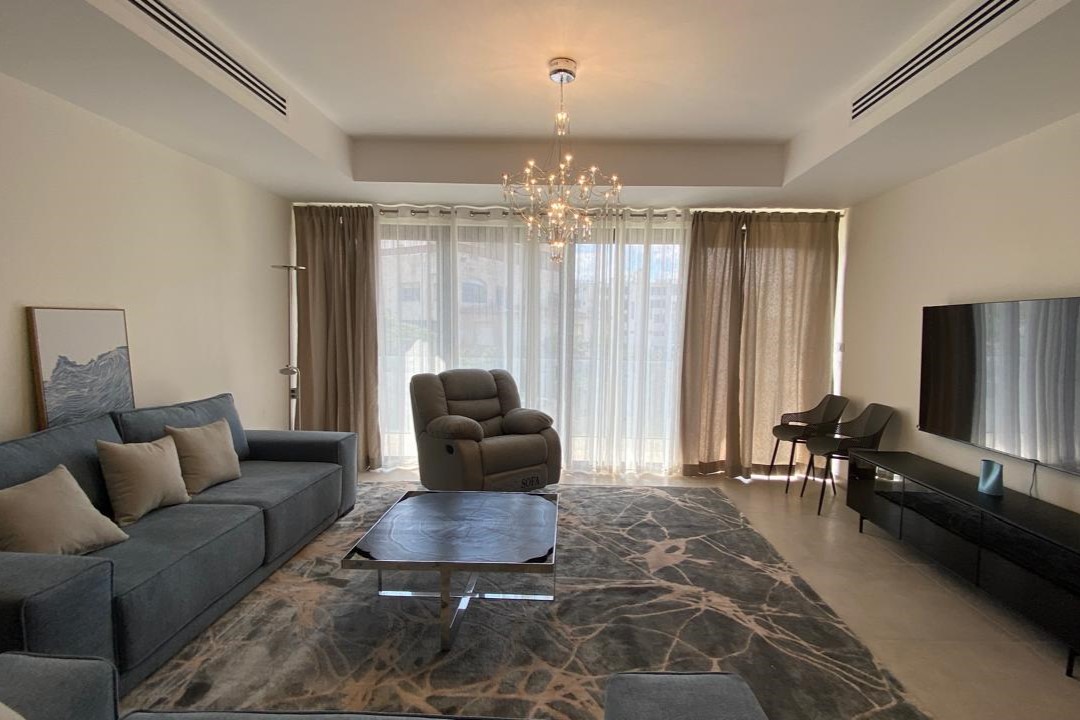Bright modern living room with a blue sofa, beige cushions, and a gray recliner facing a wall-mounted TV; large windows with sheer curtains fill the space with natural light.