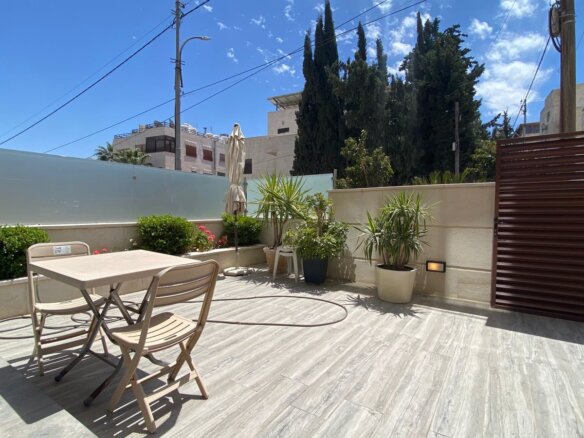 Sunlit patio with a beige plastic table and folding chairs, a closed umbrella, and potted plants along a low wall by a wooden gate.