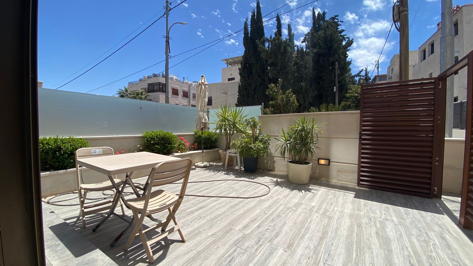 a5f6b69d-0b5e-4bbb-89bb-55f3437bbea0 Sunlit patio with a beige plastic table and folding chairs, a closed umbrella, and potted plants along a low wall by a wooden gate.