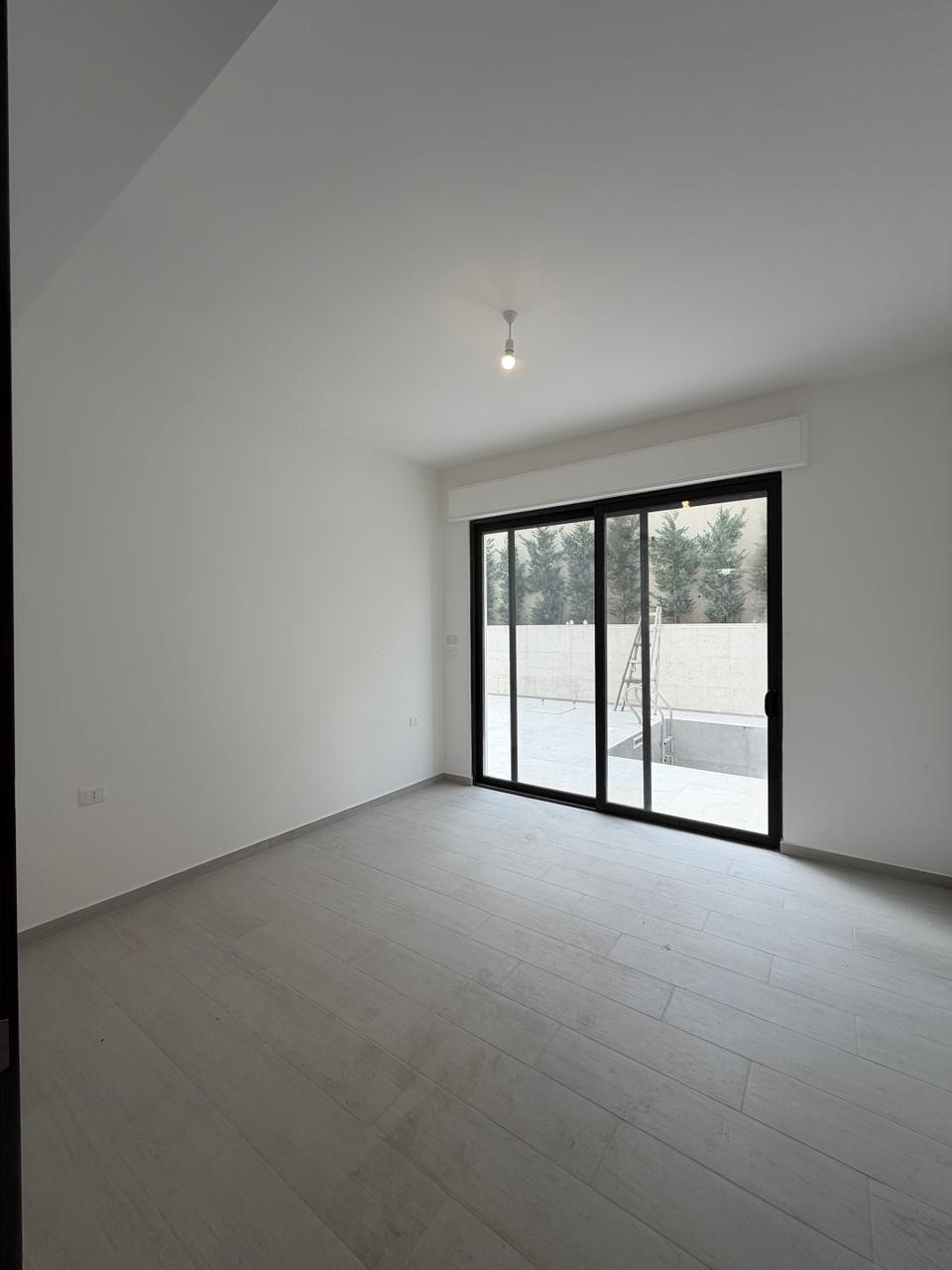 a96d40b8-9ddd-4984-aabf-99110b8a0b0e Empty modern room with white walls, light gray wood-look flooring, and a large sliding glass door opening to an outdoor area; single bare bulb ceiling light.