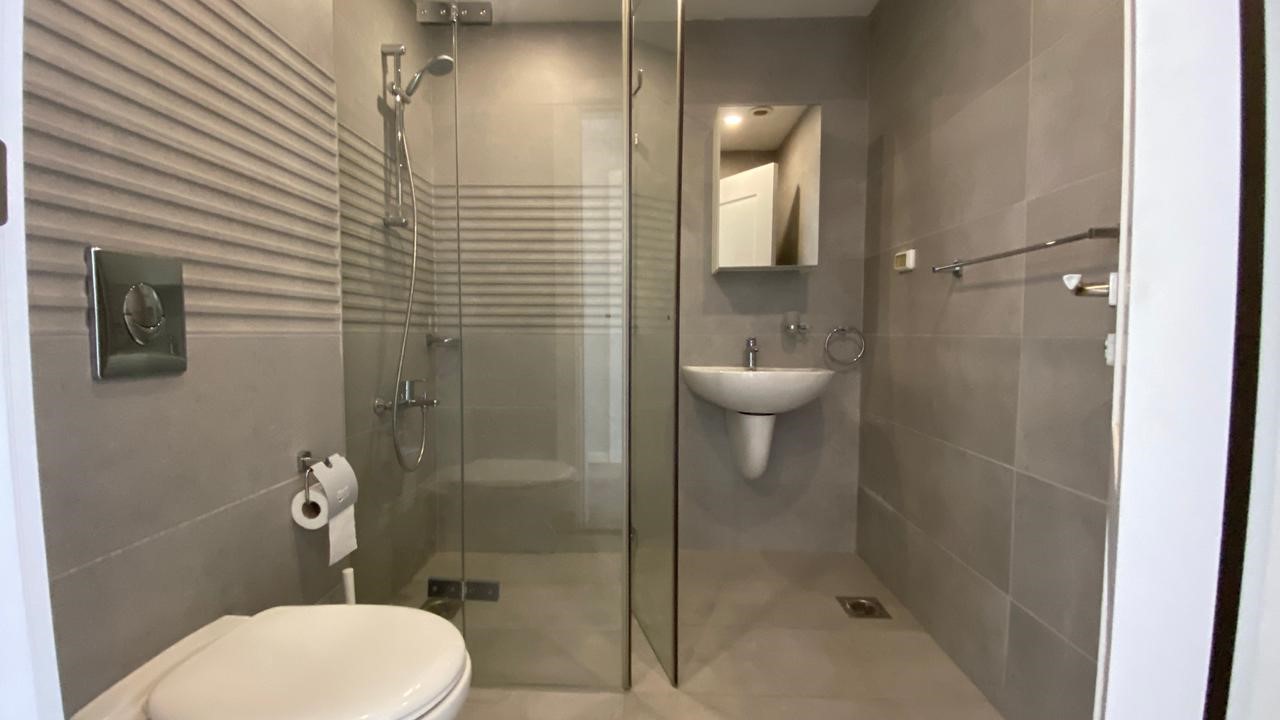 a98150a3-5dfa-476e-bbd5-b8341830b8be Modern gray-tiled bathroom with a glass shower enclosure, wall-mounted toilet, and a pedestal sink across from a mirror cabinet.