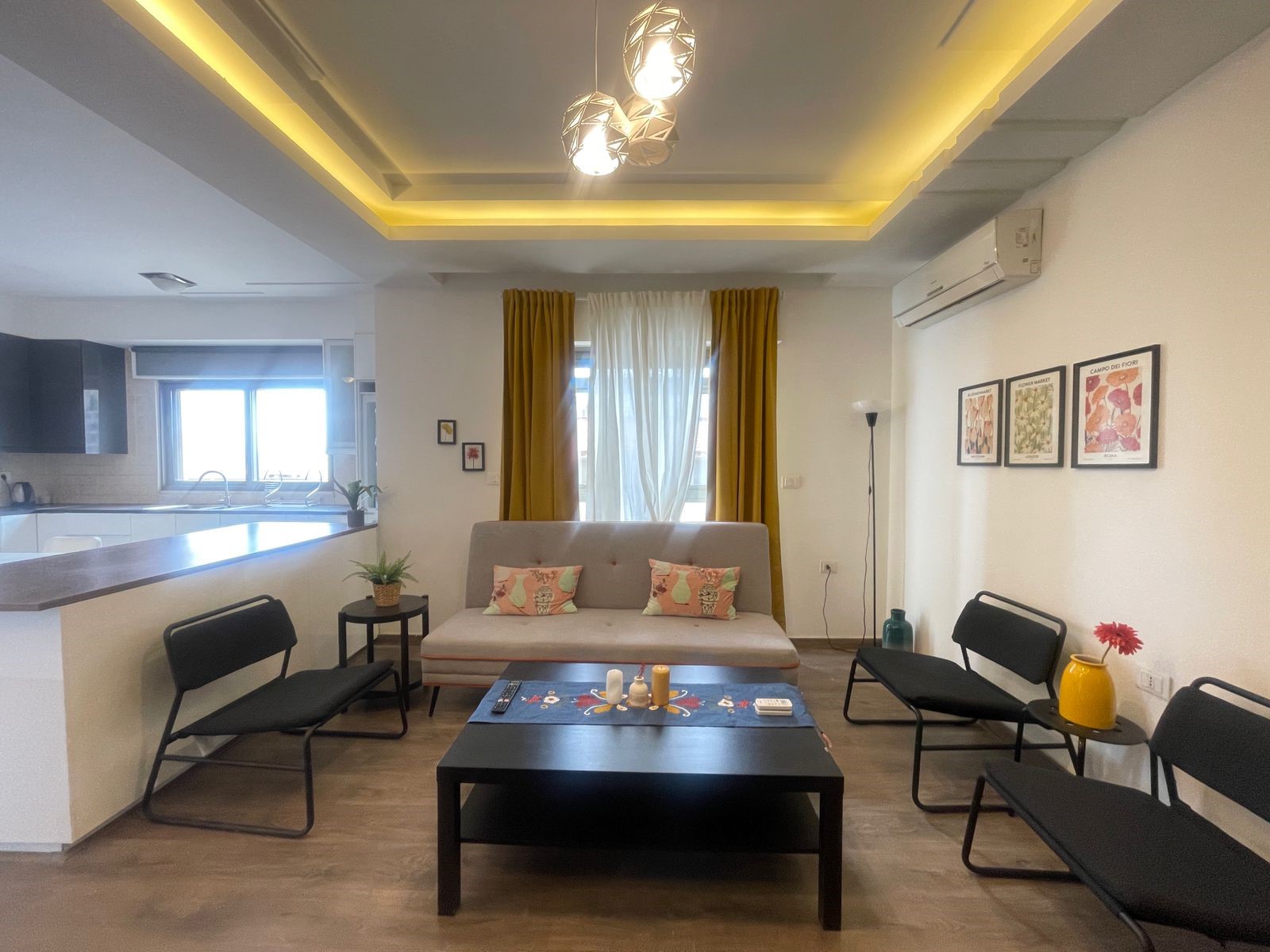 aa20c429-6389-4730-b32b-3c649f8717ce Open-plan living area with a gray sofa, tropical-pattern cushions, black coffee table, and mustard curtains framing a bright window; kitchen counter visible on the left.
