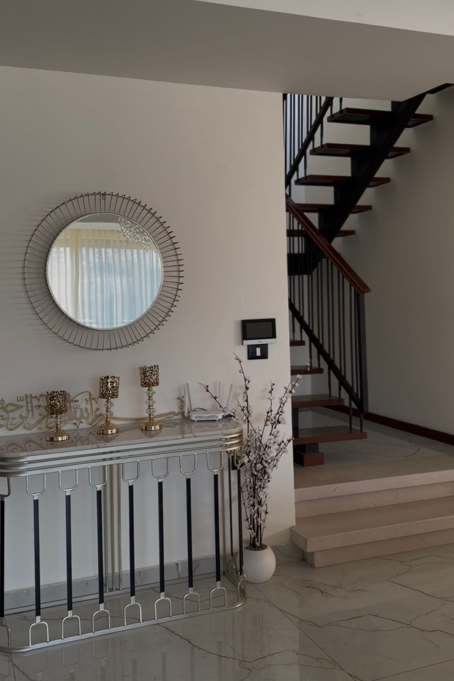 aaf6a912-42bf-4596-bc3c-23afcf264b20 Modern foyer with a round sunburst mirror above a glossy metal console and gold candle holders, against a light wall.