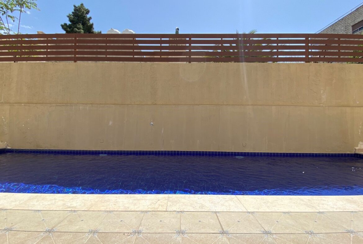 Rectangular blue-tiled pool with a tan stone deck, high beige wall, and a horizontal wooden fence on a sunny day.