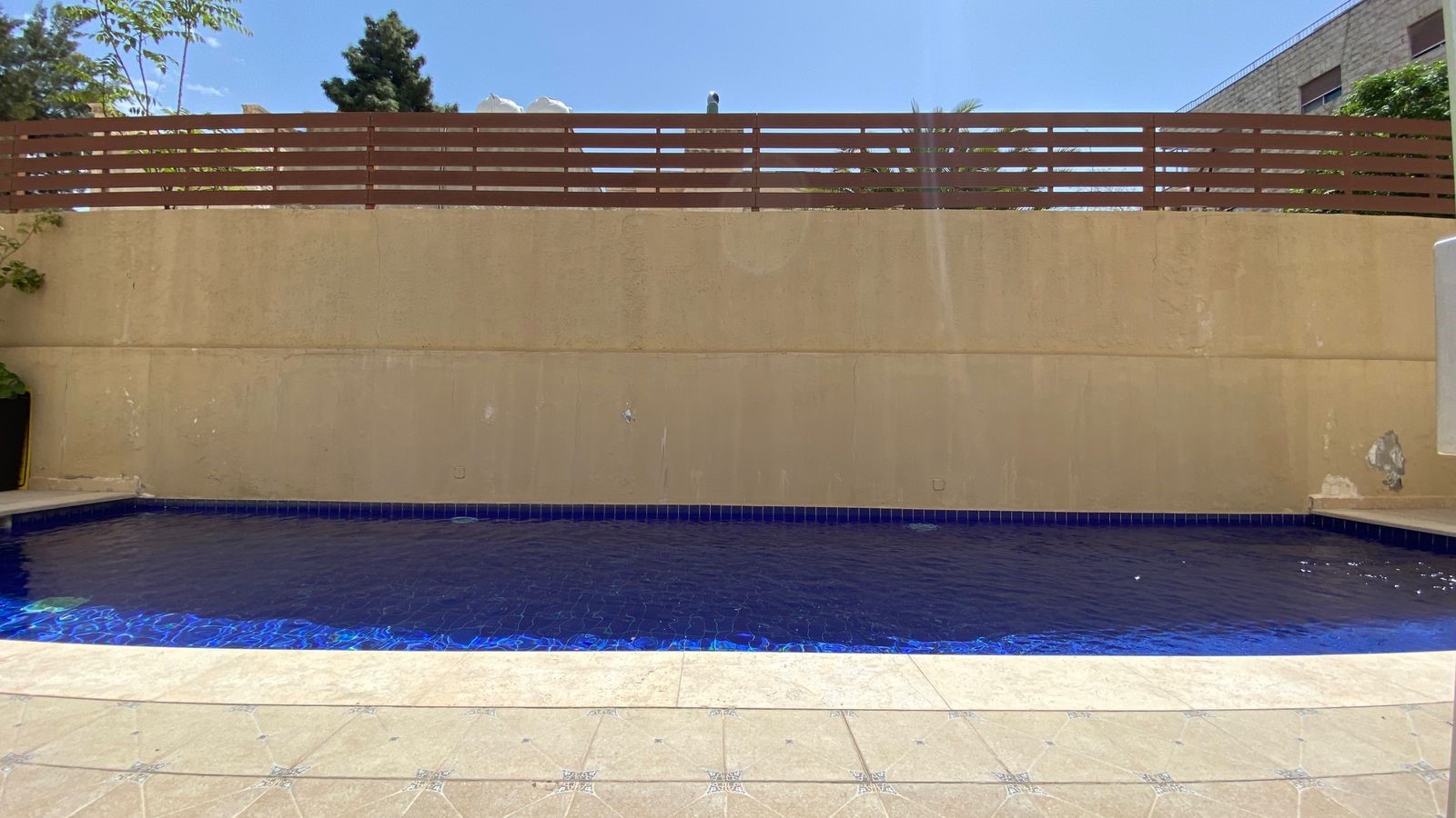 abc08dc6-be4e-4da2-ad16-679ea3a81472 Rectangular blue-tiled pool with a tan stone deck, high beige wall, and a horizontal wooden fence on a sunny day.