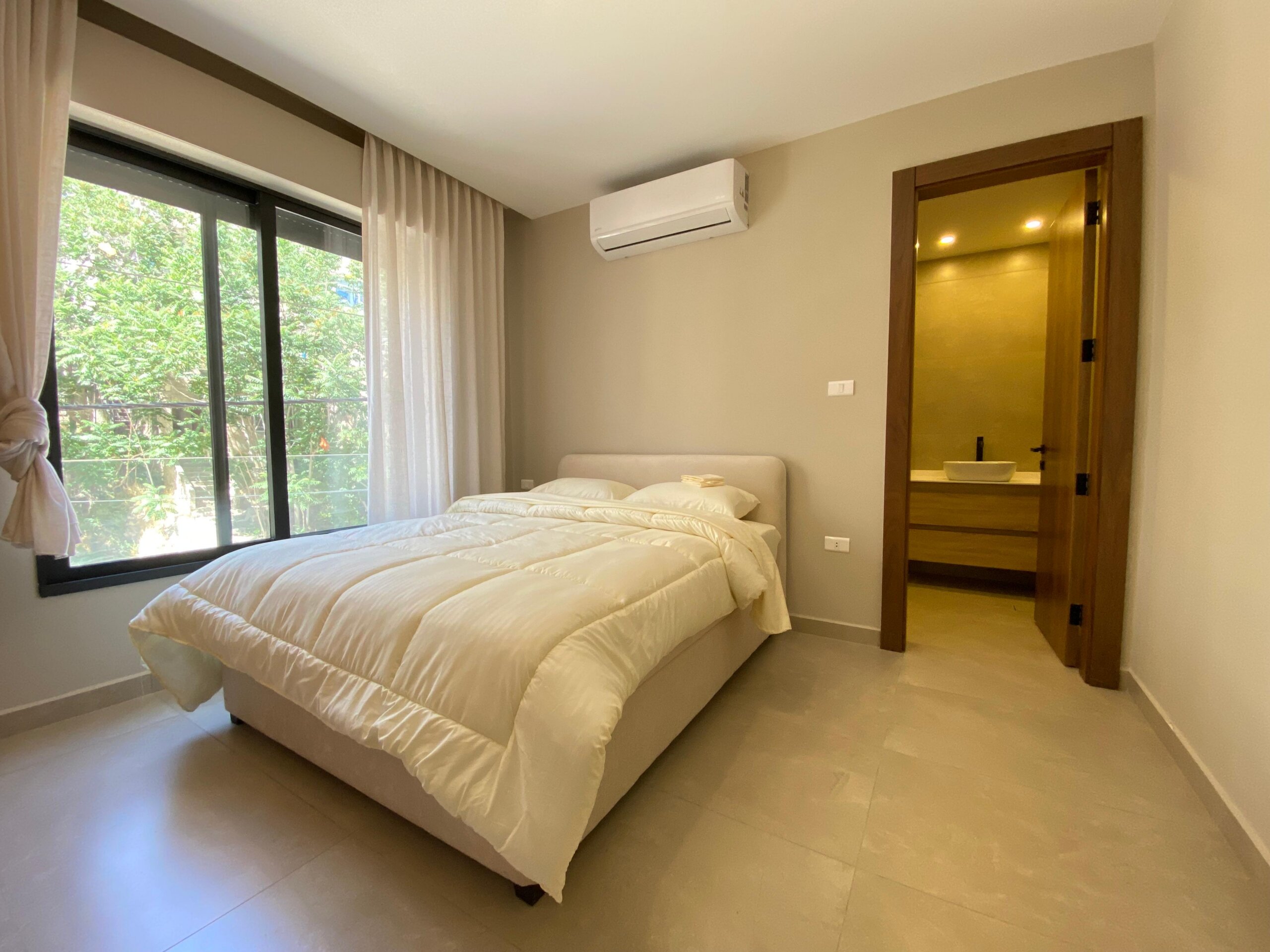ac4ced41-eb80-41e8-989e-2cc7c5b530fa Bright hotel-style bedroom with a white comforter on a bed, beige walls, large window with sheer curtains, and a wall-mounted air conditioner above-side.