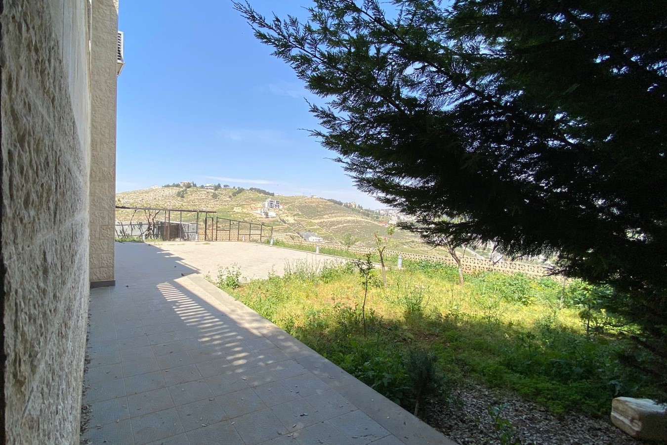 ac6bd831-543f-4292-b90b-391a6ff7432a Sunlit terrace beside a stone wall, overlooking terraced hills and vineyards under a clear blue sky, with a large tree providing shade on the right.