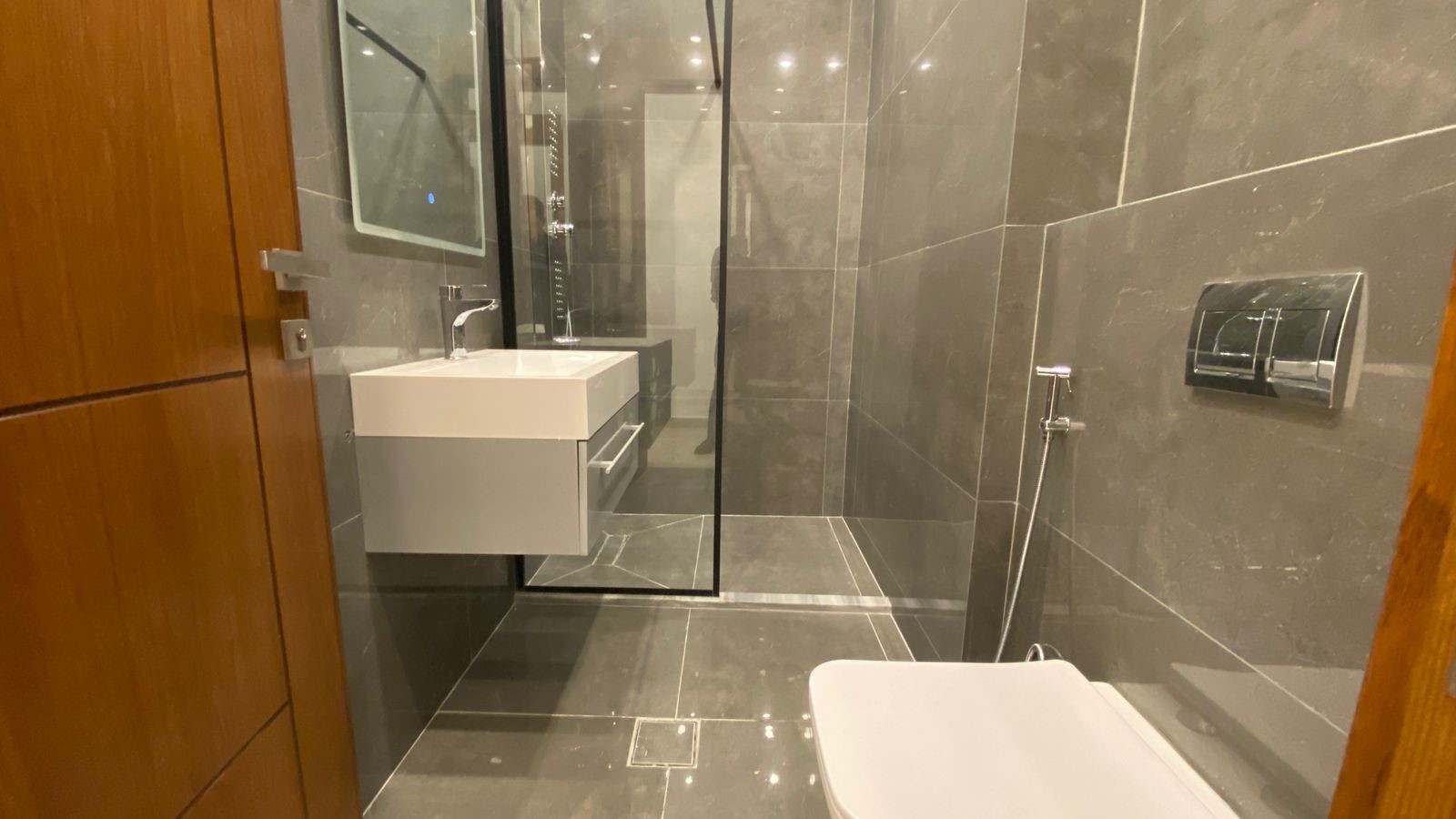 ac80125a-6e34-4ea1-8cdf-3b6b8351486d Open wooden door leads into a modern gray-tiled bathroom with a glass shower enclosure, wall-mounted sink, and toilet area.