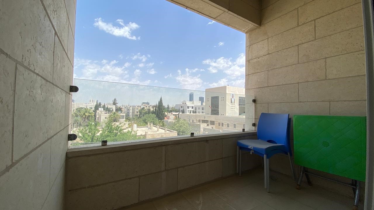 acaaf48c-8d9c-4694-acfb-c01af6a27453 Balcony with glass railing overlooking a cityscape and blue sky; blue chair and green foldable table against beige brick wall.