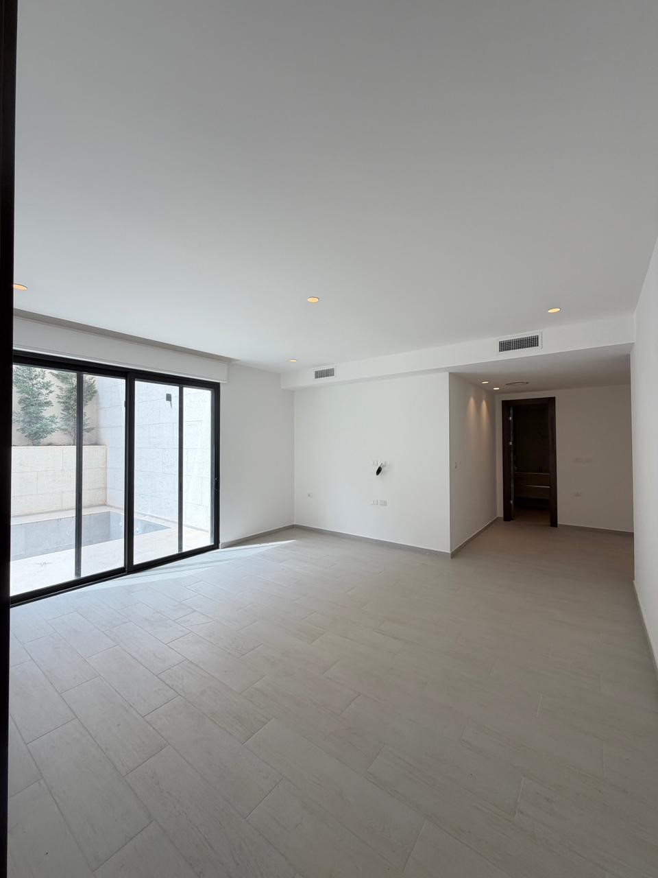 ae0994e8-cc19-4711-b209-ad466eec463c Empty modern living room with large sliding glass doors opening to a patio, light gray tile floor, and white walls with recessed ceiling lights on.