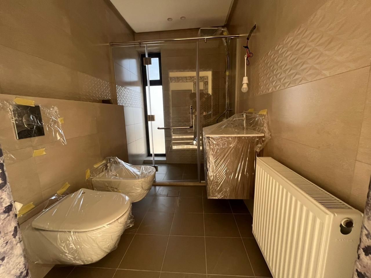 ae8a251f-de23-4c58-9a11-eee4e3c3469a Newly tiled bathroom under construction with wrapped toilet and bidet, glass shower, wall sink, and radiator.