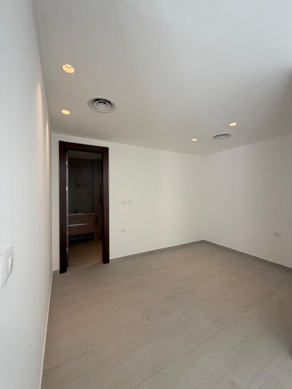 aee6adb2-c9d0-441c-b9ba-0747dde8f37b Empty modern room with white walls, light gray floor tiles, recessed ceiling lights, and a doorway to a bathroom.