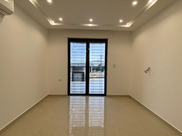 Empty rectangular room with glossy light tile floor, white walls, and recessed ceiling lights; left wall has a wall-mounted air conditioner, and at the far end are barred glass double doors opening to a balcony.