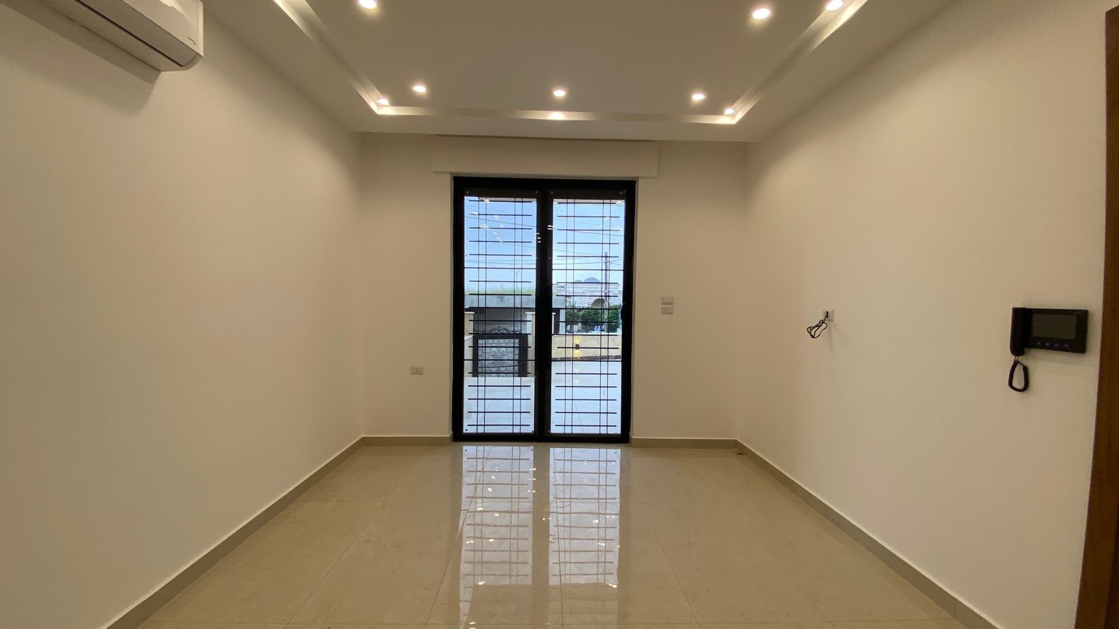 af49c16a-3d2f-4a4d-9198-30d01f348fbf Empty rectangular room with glossy light tile floor, white walls, and recessed ceiling lights; left wall has a wall-mounted air conditioner, and at the far end are barred glass double doors opening to a balcony.