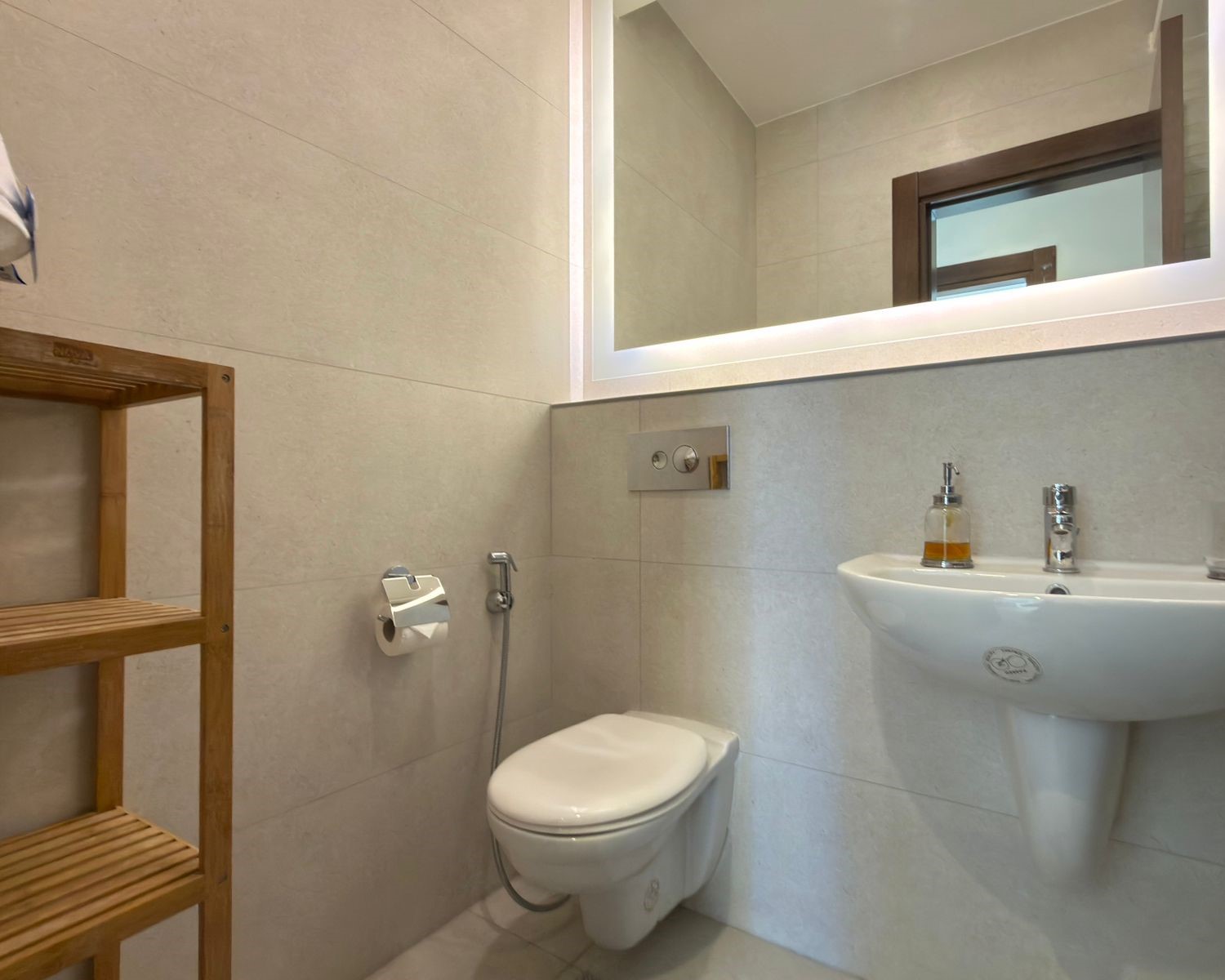 af815c42-8b8b-4398-be67-054099c2535a Modern bathroom with wall-mounted toilet, white sink, and wooden open shelf; backlit mirror above the sink to the right.