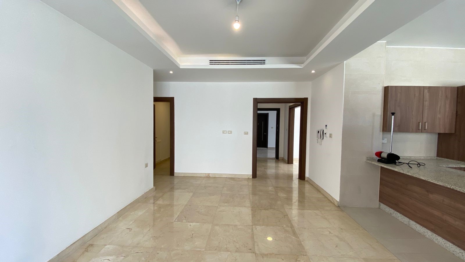 affa5f54-dfbd-4a65-ae79-6d59fe7a59a6 Bright empty open-plan living area with marble tile floor, white walls, and a kitchen on the right; a mop stands by the counter.
