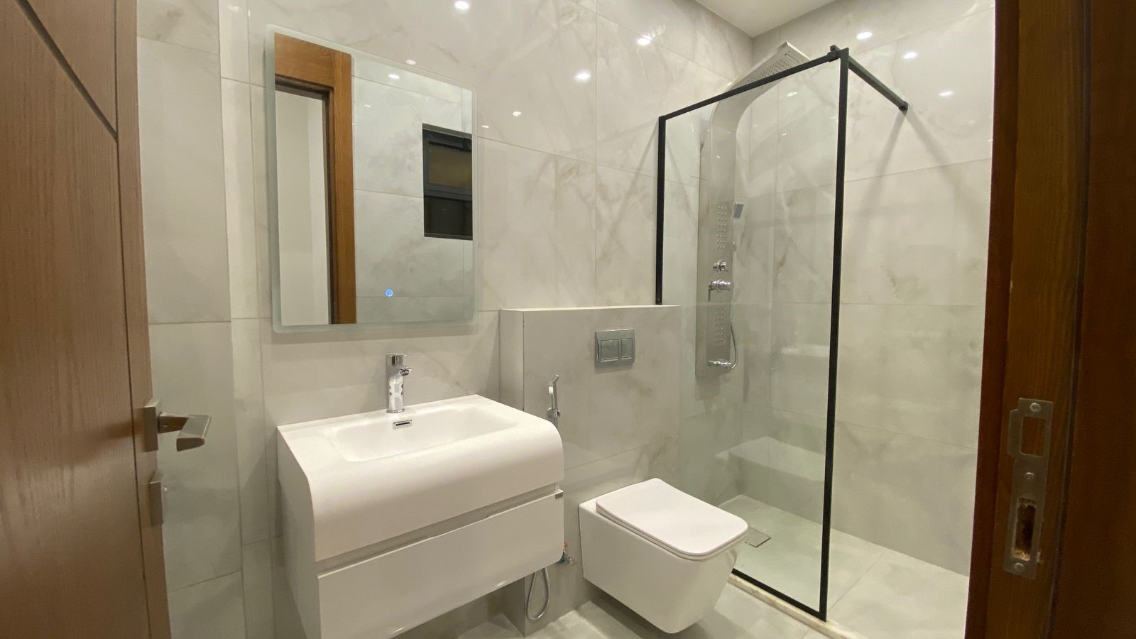 b1b9fefc-e281-4dac-b7c0-f0ff57a7a84d Modern white bathroom with a wall-mounted sink, wall-hung toilet, and glass-framed shower on marble tile walls.