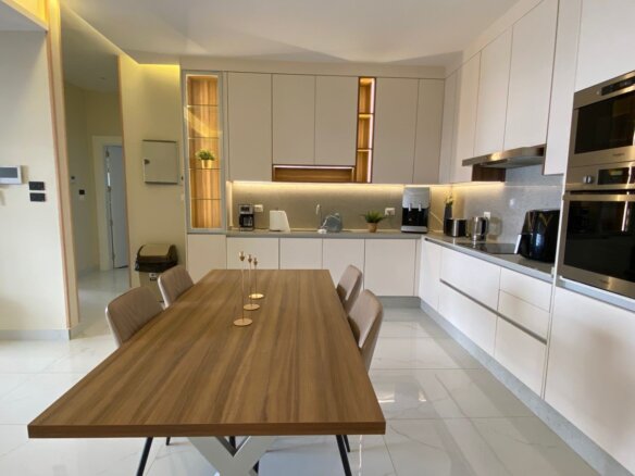 Modern kitchen with a long wooden dining table and six upholstered chairs, white cabinets, and built-in stainless steel ovens on the right.