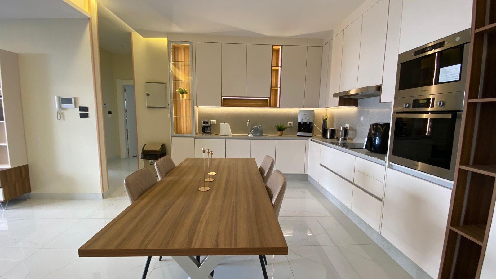 b252af50-83f0-4d2b-9d23-8555136a7388 Modern kitchen with a long wooden dining table and six upholstered chairs, white cabinets, and built-in stainless steel ovens on the right.