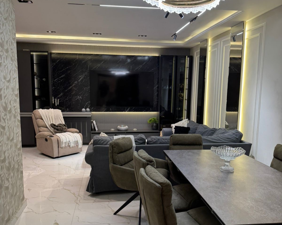 b2eac646-9733-435a-be9c-81a6583b796e Luxurious living room featuring a large wall-mounted TV on black marble, a gray sectional sofa, and a beige recliner with a blanket.