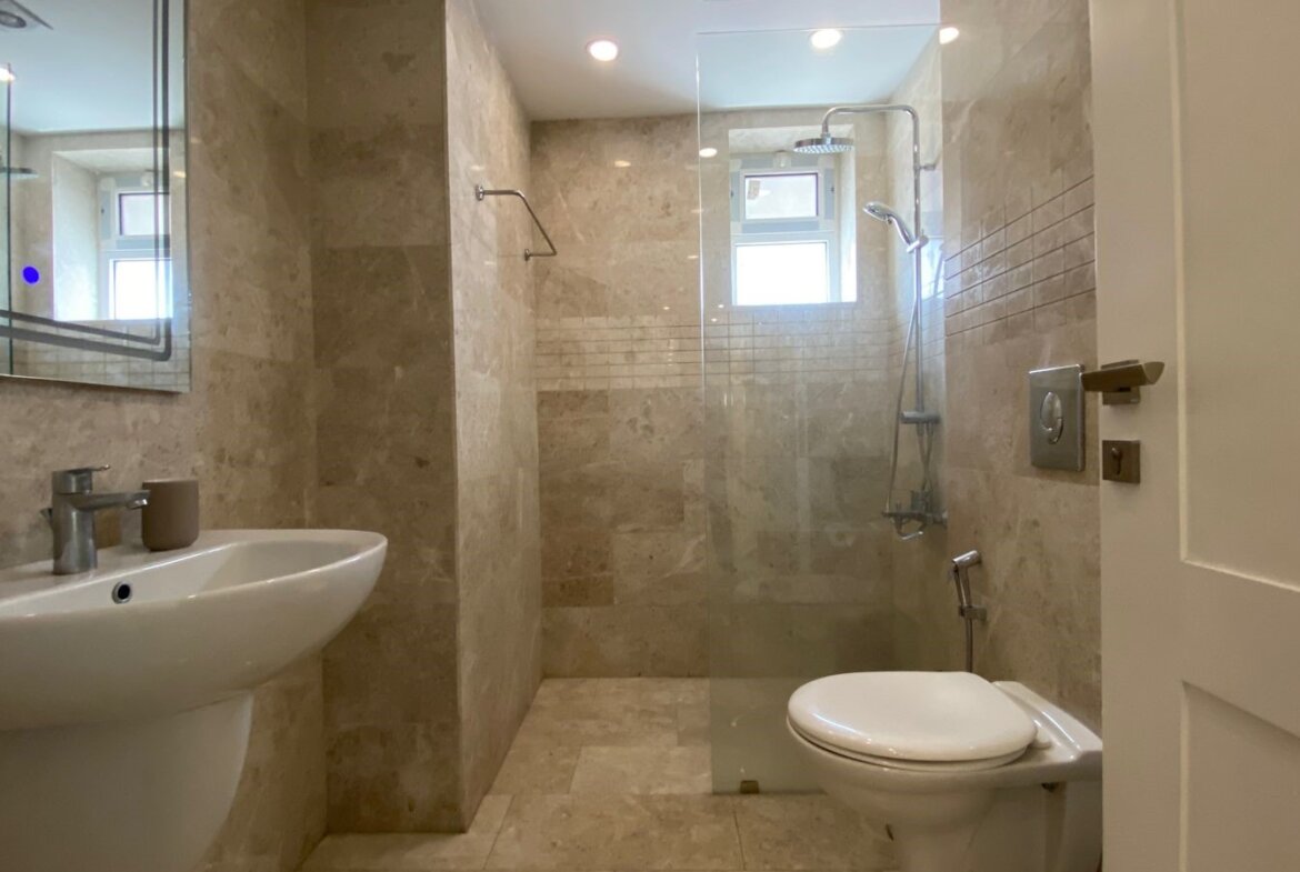 Contemporary bathroom with beige marble tiles, a white pedestal sink, and a glass-enclosed shower area opposite a suspended toilet.