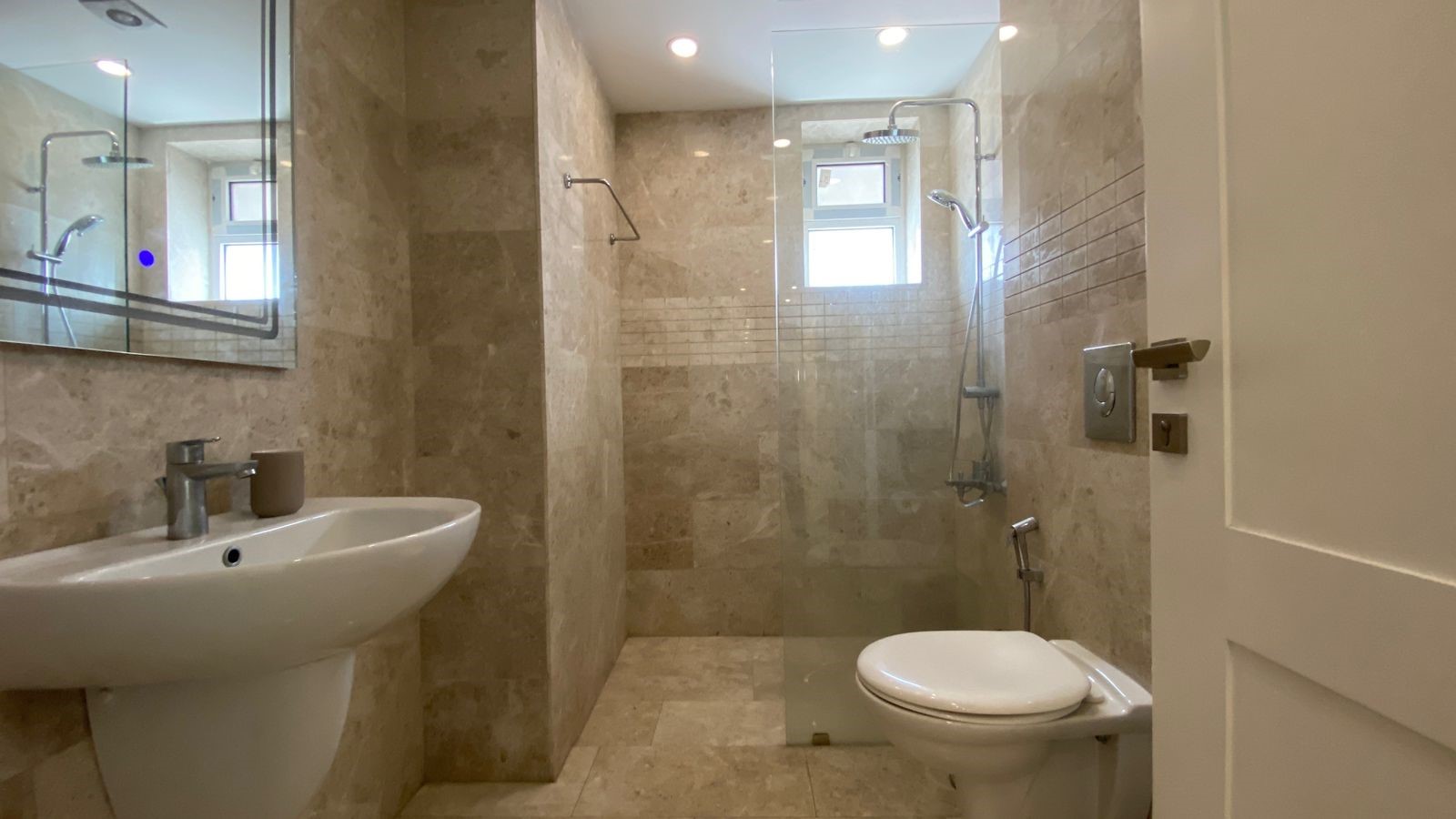 b35d5290-f686-4e6b-8b0a-3d92263c7eb7 Contemporary bathroom with beige marble tiles, a white pedestal sink, and a glass-enclosed shower area opposite a suspended toilet.