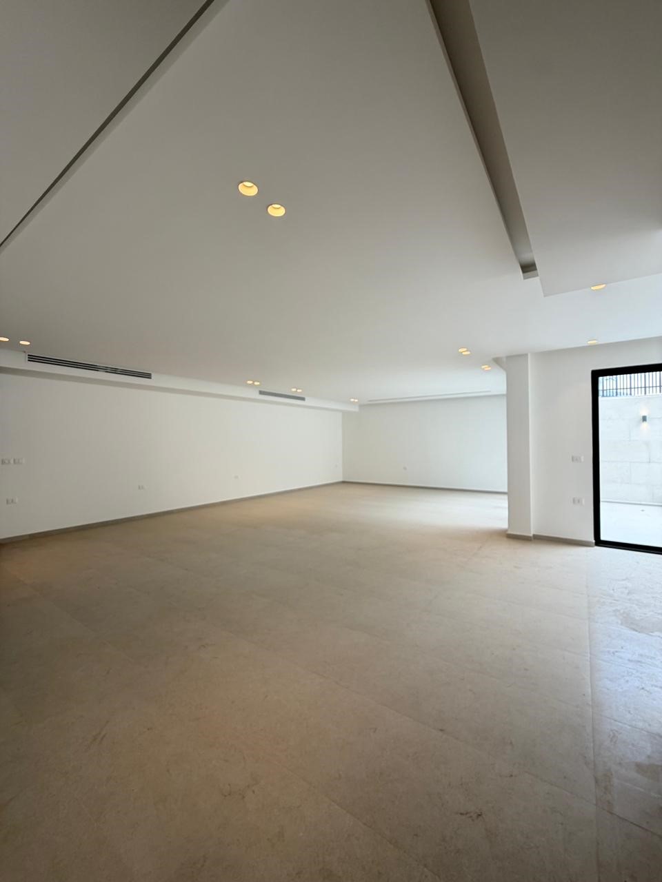b3857631-d89c-490c-9427-dbe5c528f846 Empty, sunlit white-walled gallery or showroom with beige tile floor, recessed ceiling lights, and a large glass door on the right.