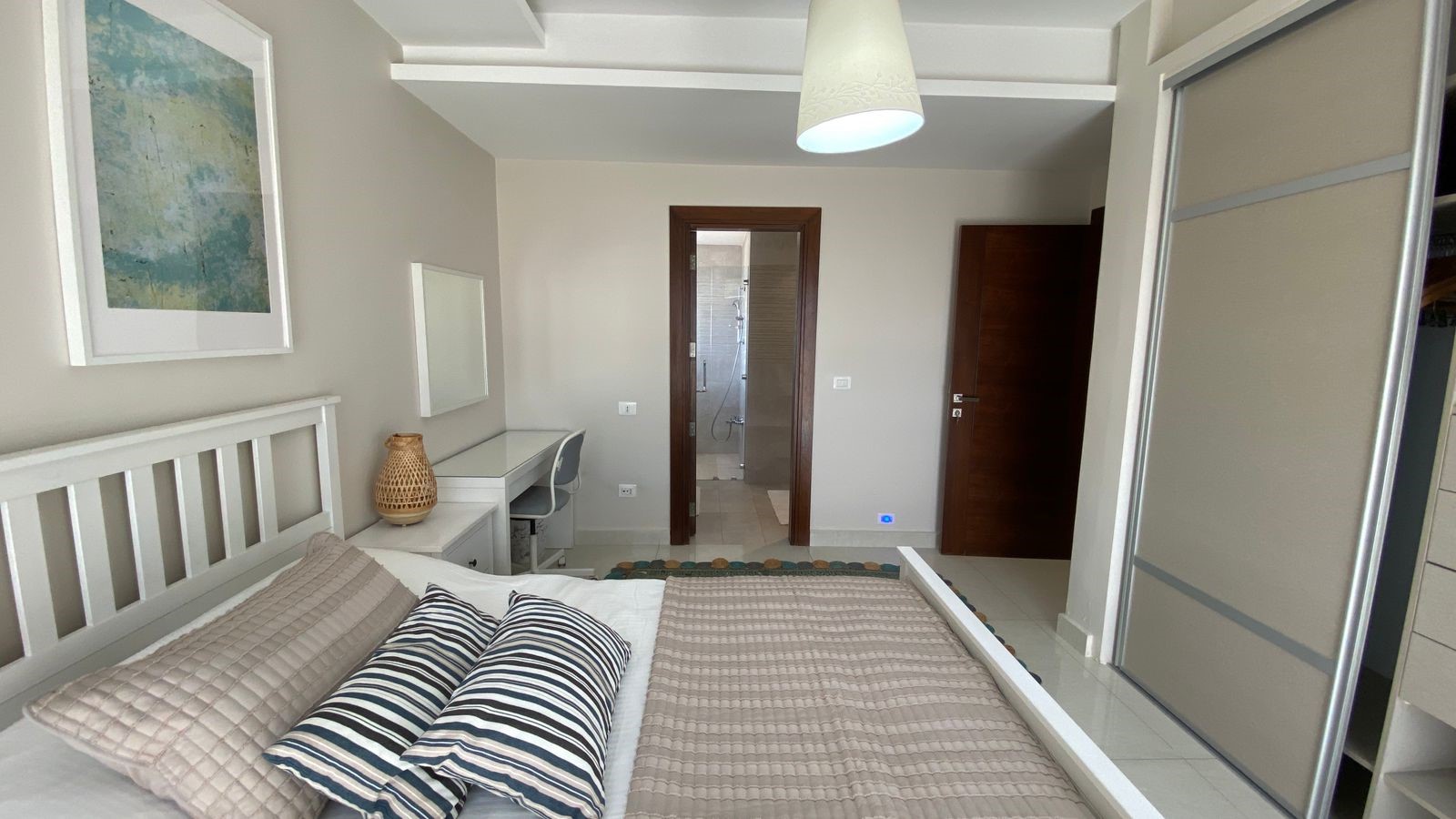 b38d9187-371f-4245-9270-0301346e7197 White wooden bed with beige quilt and striped pillows in a bright gray bedroom; a desk and chair against the far wall, framed art above the headboard, and an open doorway to a glass-walled bathroom.