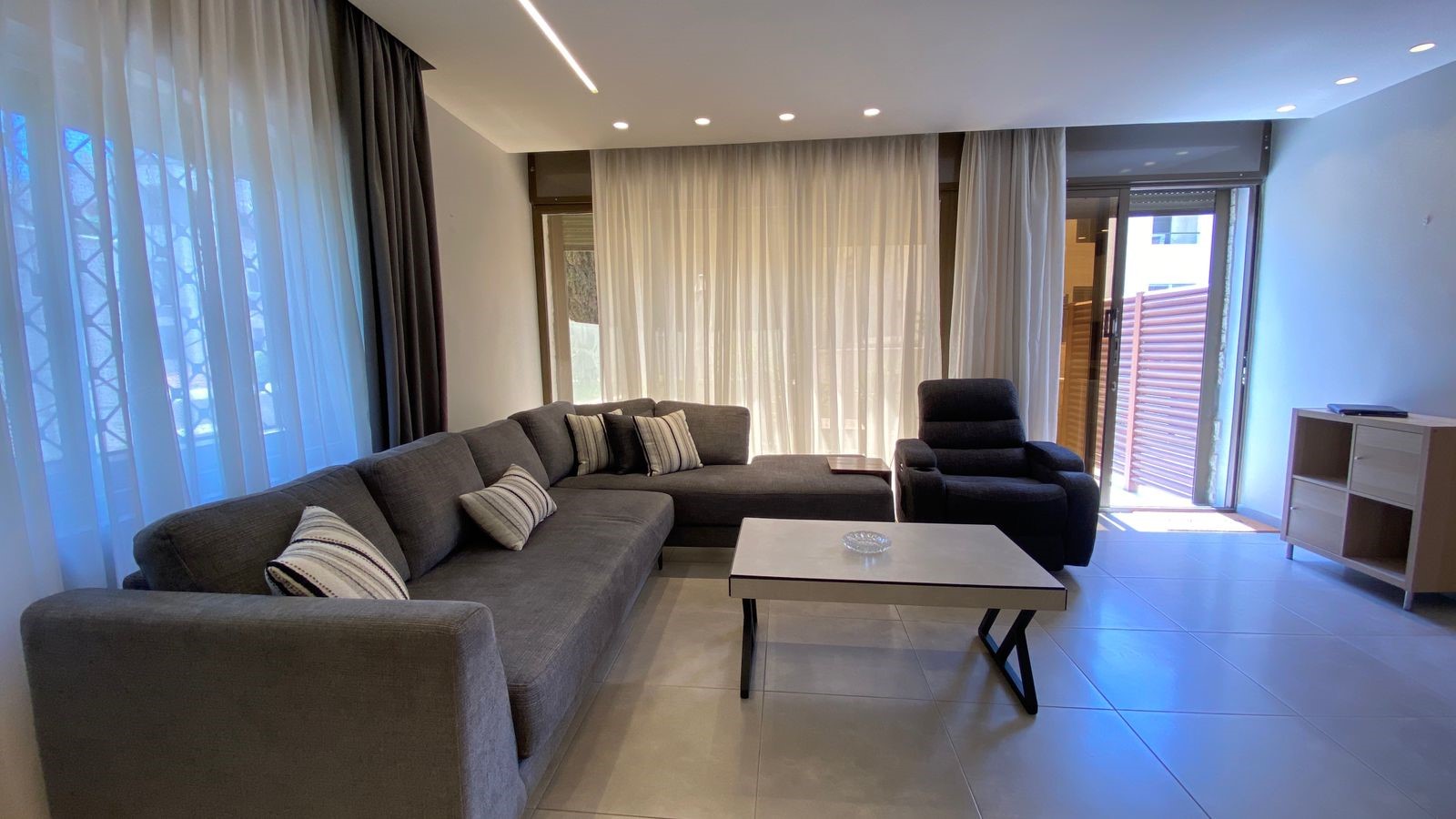 b454a0b5-a879-417a-b203-09131352485f Modern living room with a gray L-shaped sofa, striped cushions, and a black recliner around a rectangular coffee table.