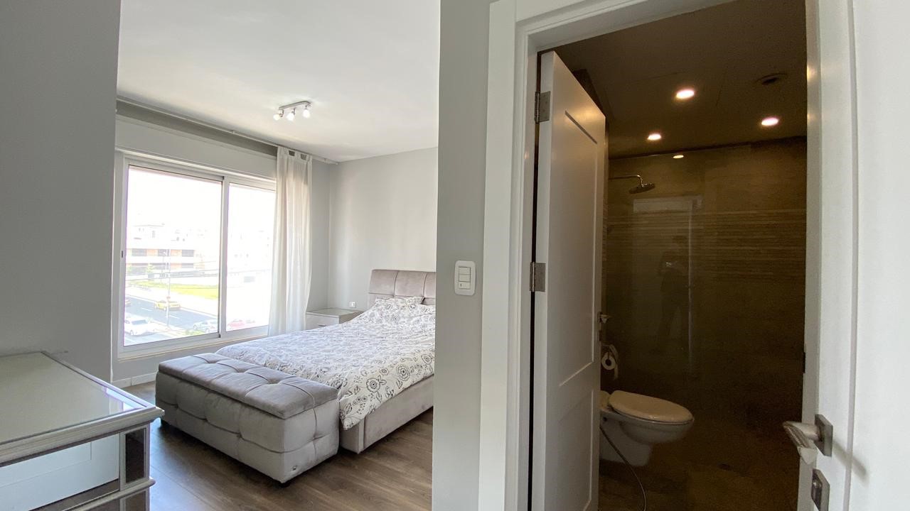 b564d2c9-d1bf-47c7-80b6-74d3e42e639c Bright bedroom with a large window, queen bed, gray upholstered headboard, and a matching bench at the foot near white nightstands and sheer curtains. Two-tone flooring visible. Bedroom with large window view, upholstered bed, and a white dresser; doorway opens to a dark, glass-enclosed bathroom across from the bed.