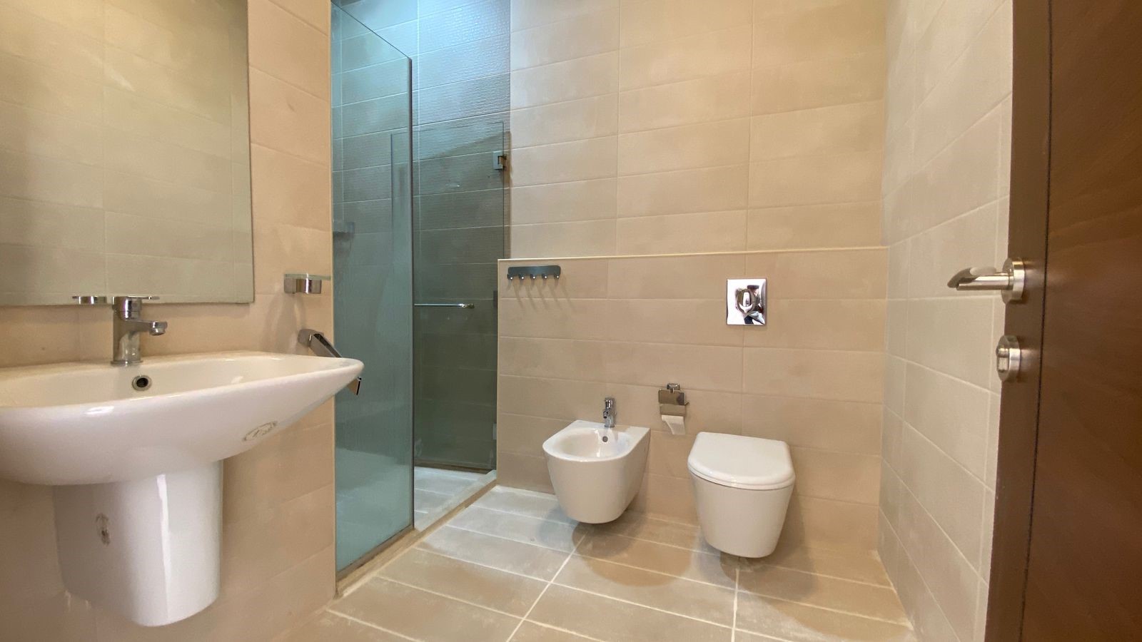 b5694252-46c2-48cf-bf94-3a68d9d3be40 Beige-tiled bathroom with a white pedestal sink, glass shower enclosure, bidet, and a wall-mounted toilet near the doorway.