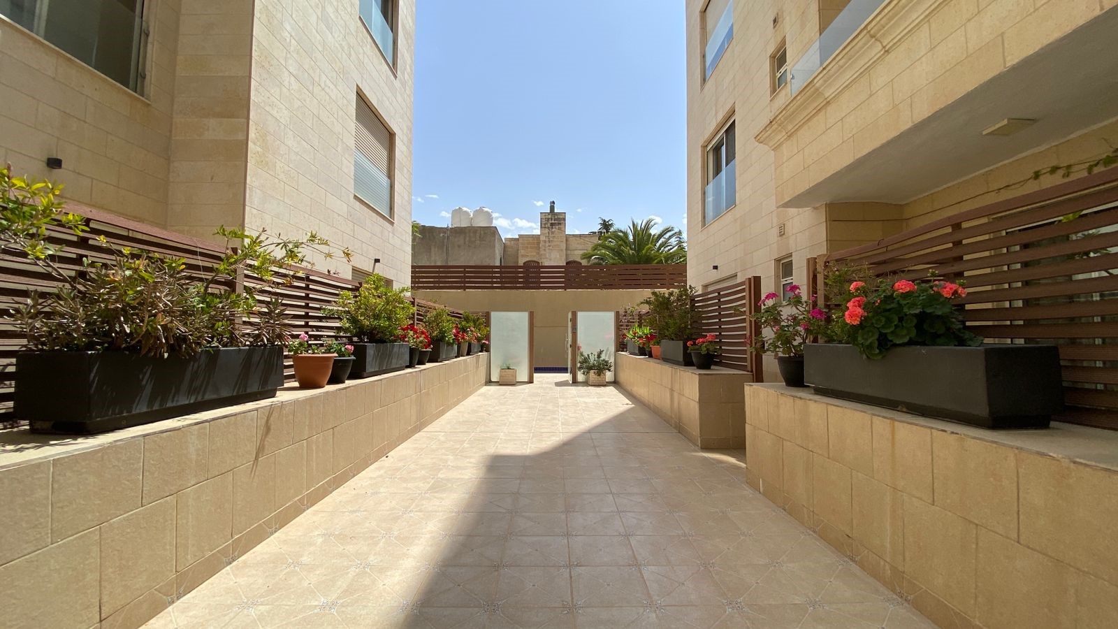 b70ff406-3d10-46b5-817a-c471f4ee033b Sunlit courtyard between beige stone apartment buildings with a row of potted plants on low walls and a wooden fence on both sides.