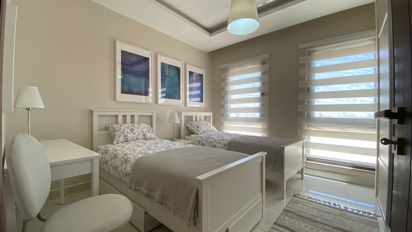 b7727d89-0d66-4add-8223-5534f06fdd5d Twin white beds with floral grey bedding in a bright, modern bedroom with matching nightstands and lamps.