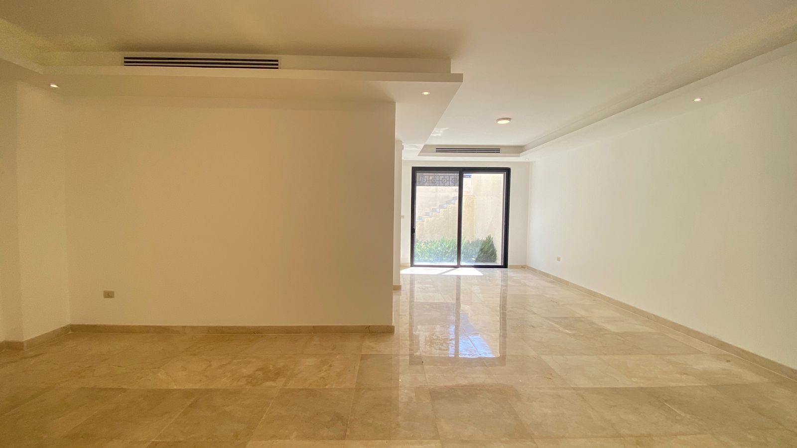 b8cc2714-3ffb-432b-a461-fcdcf1b2e39a Bright empty living room with beige marble flooring, white walls, recessed ceiling lights, and a glass sliding door to an outdoor area.