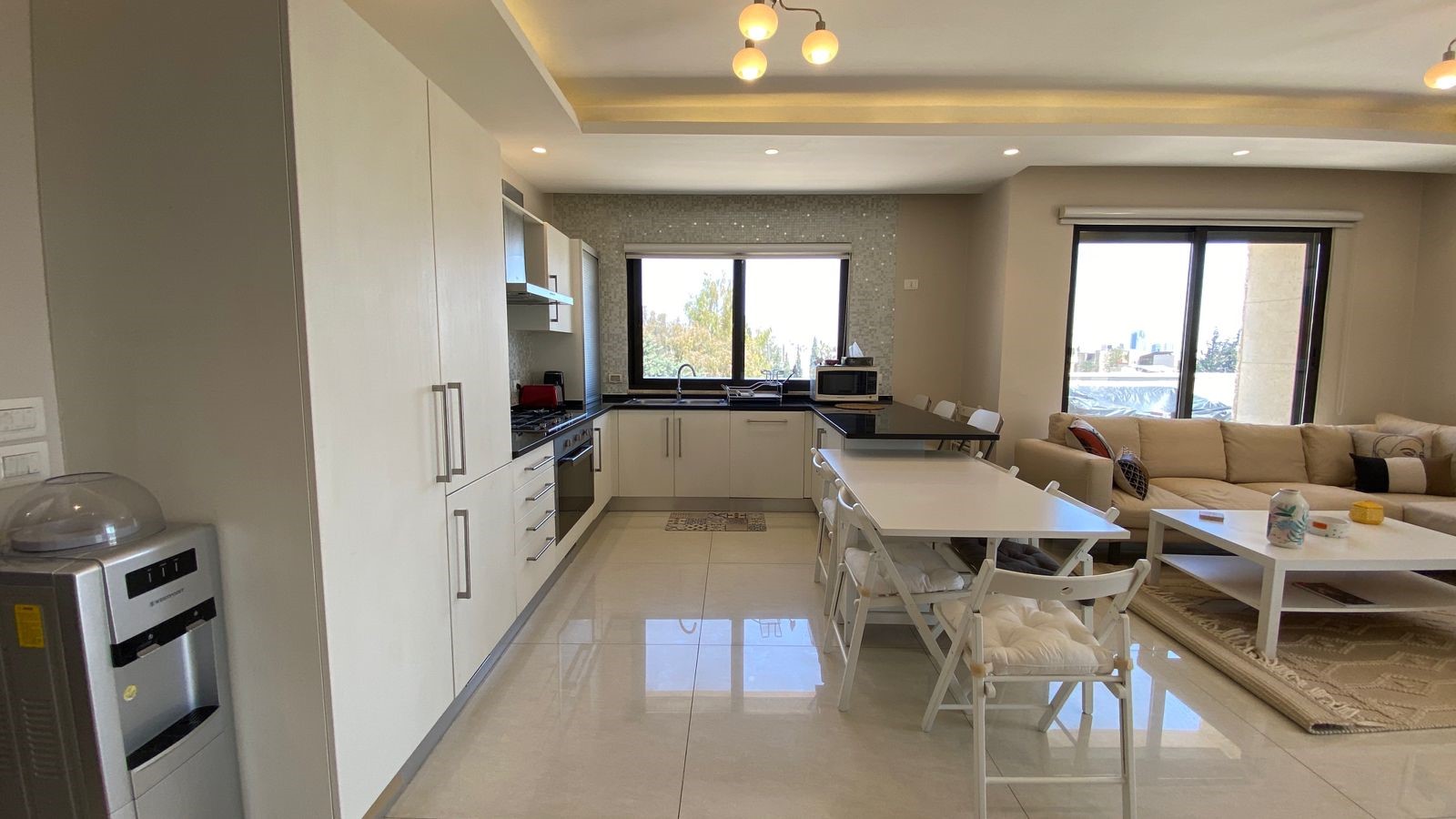 b927c2f3-5c45-420a-94e7-6dddc67f2327 Open-plan kitchen and living room with white cabinets, an island, and a beige sectional by large windows.