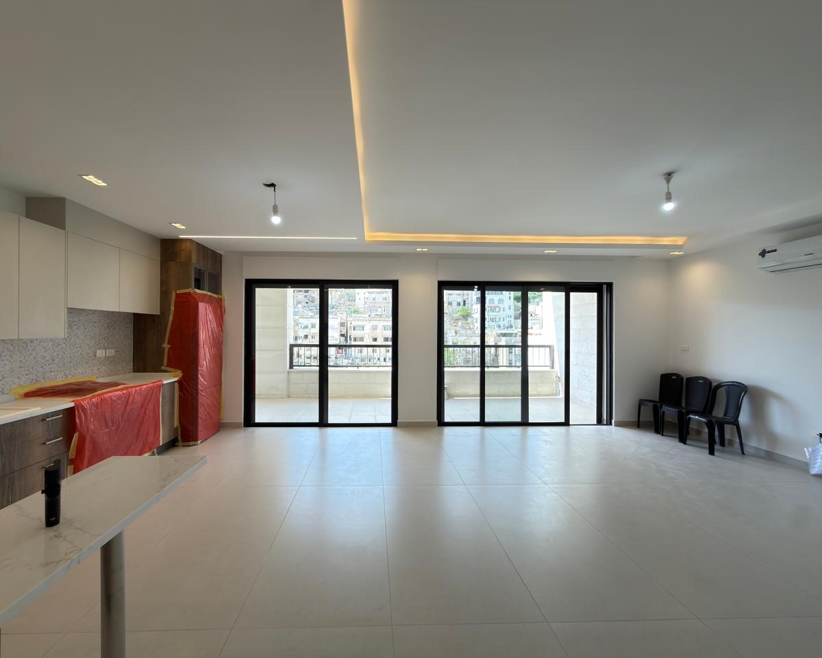 b9803c2e-d678-4131-81a5-31d9590adb1f Empty modern living area with a kitchen to the left, red protective coverings, and three black chairs by the right wall.