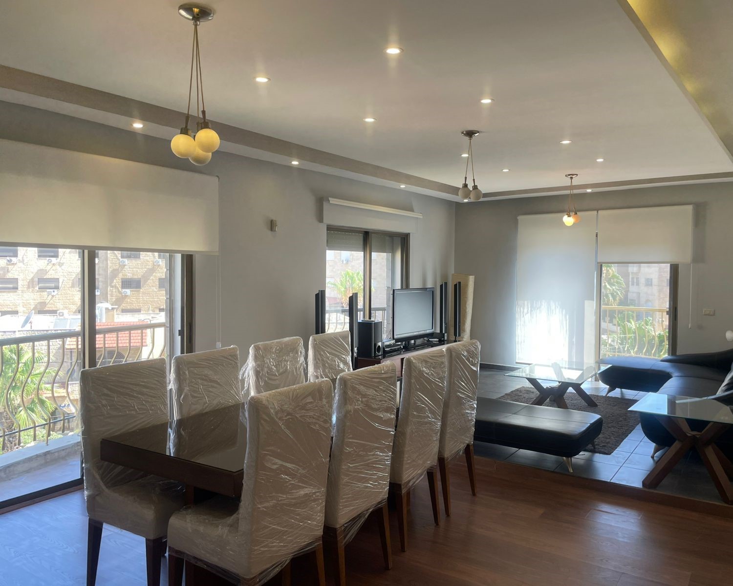 babd0540-a0fe-478e-a29f-35f929ae7123 Open‑plan living and dining area with a dark wood table and white plastic‑wrapped chairs, a TV on a stand, and sliding balcony doors in view.