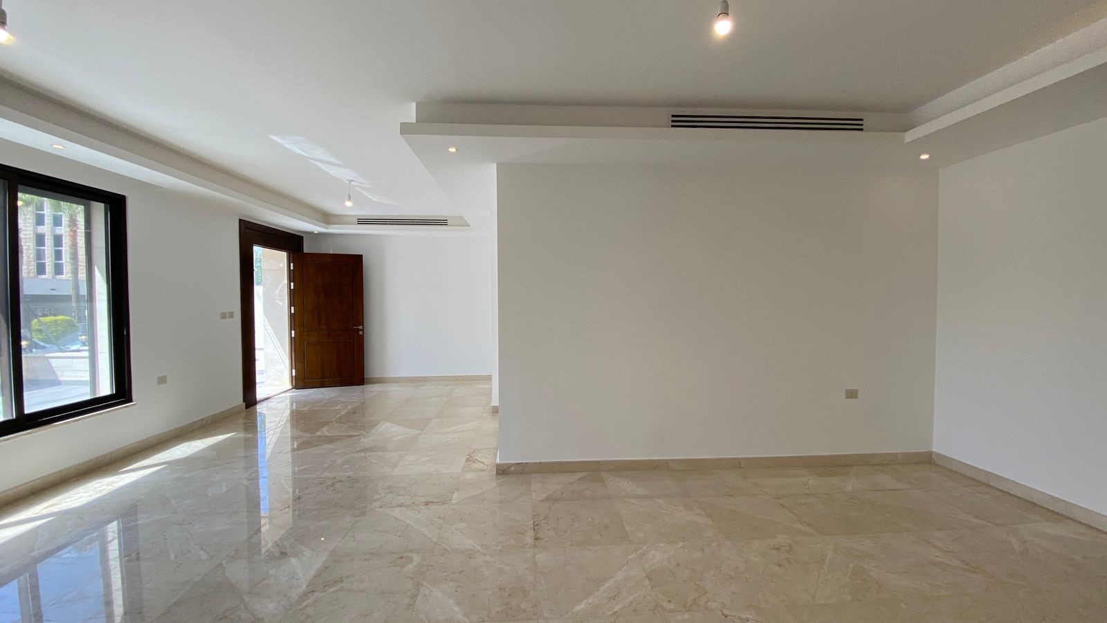 bb166845-c397-43bd-9111-af9f491b5634 Spacious empty living area with marble floors, white walls, large window on left, and a wooden door open to the outside; ceiling track lights and vents visible.