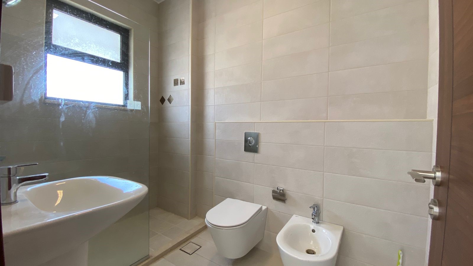 bd4e8d4b-d008-4668-8d25-6f6d1ef1db95 Modern beige-tiled bathroom with wall-mounted toilet, bidet, white sink, and a glass-enclosed shower; frosted window on the left.
