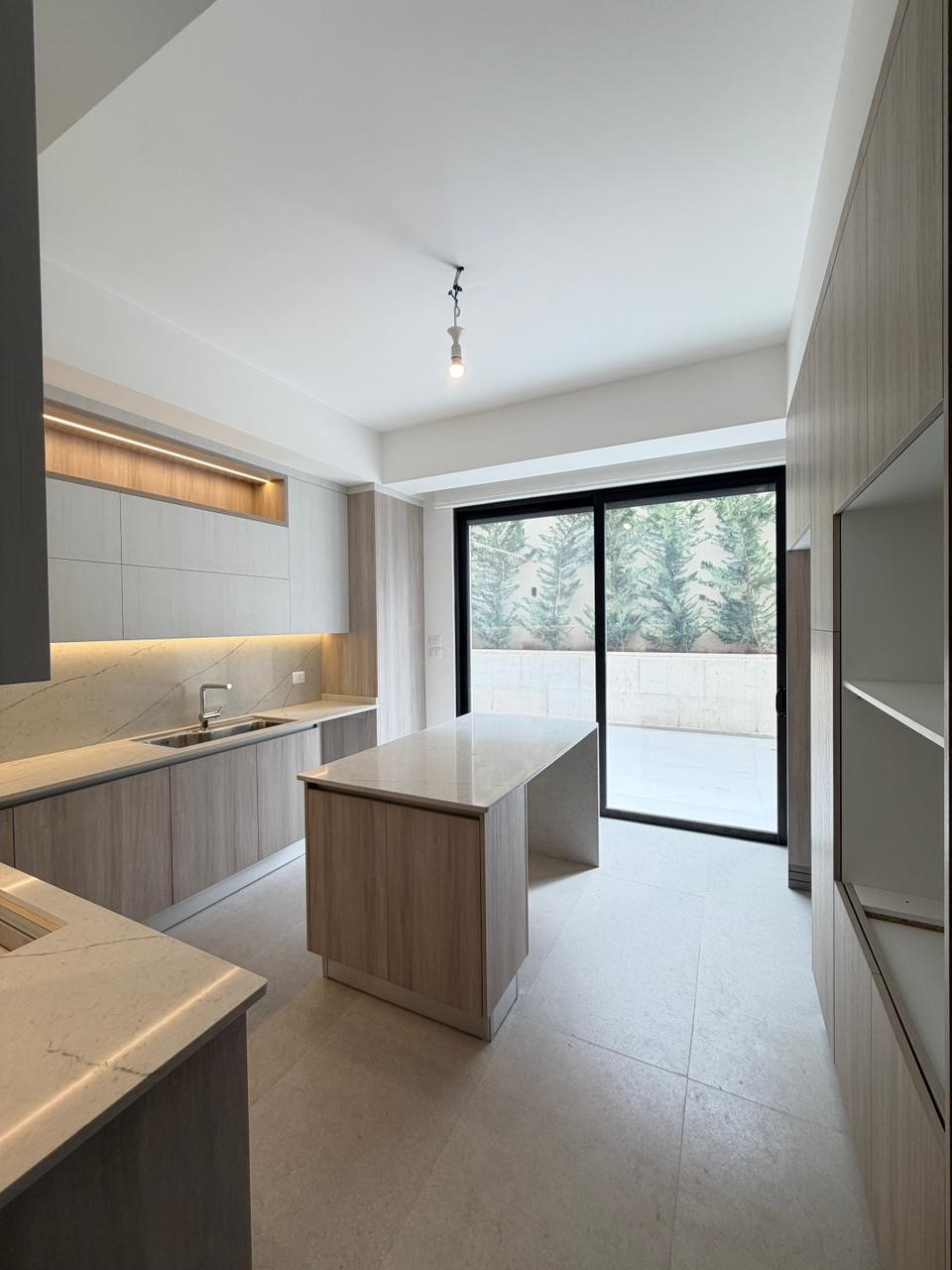 bdf5c012-2ff9-4fb4-ae54-c414b66a9912 Modern kitchen with light wood cabinets, marble countertops, and a central island; large sliding door opens to an outdoor area with a stone wall and trees outside.