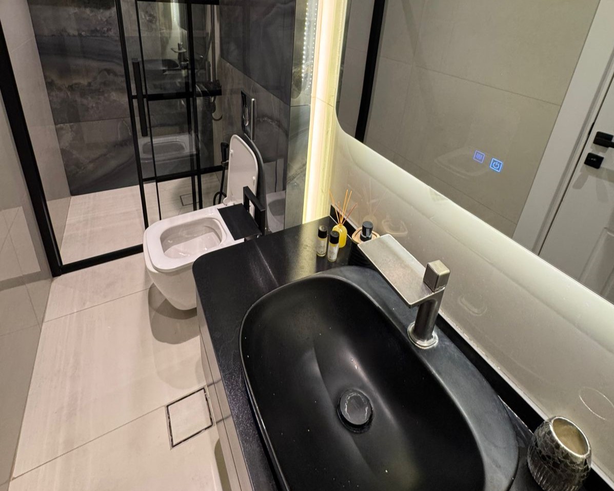 be6ef790-9a33-48ae-a16a-798d9b03be70 Modern bathroom with a black oval sink on a dark countertop, silver faucet, and a glass-walled shower in the background; toilet to the left full view shows open lid, and toiletries on the counter nearby.
