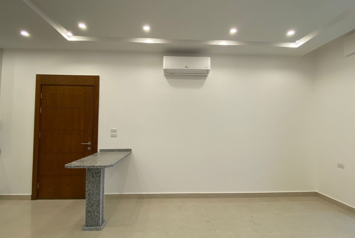 Empty modern room with white walls, a wooden door, granite counter island, ceiling spotlights, and a wall-mounted AC unit.