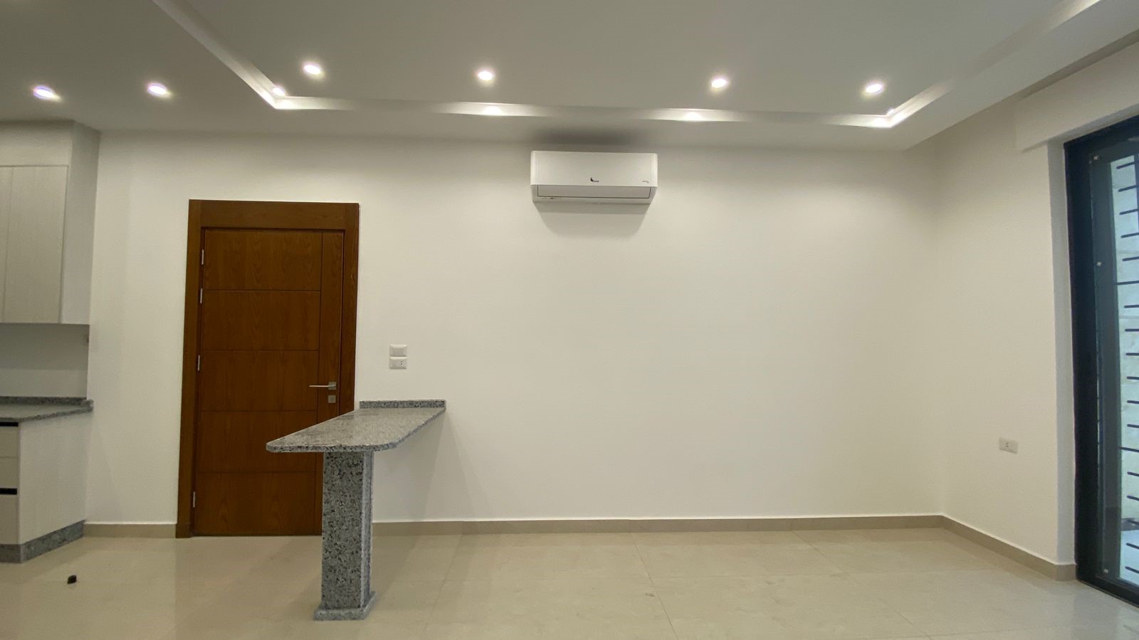 bed195a1-2ab4-45de-b994-a7c562d363da Empty modern room with white walls, a wooden door, granite counter island, ceiling spotlights, and a wall-mounted AC unit.