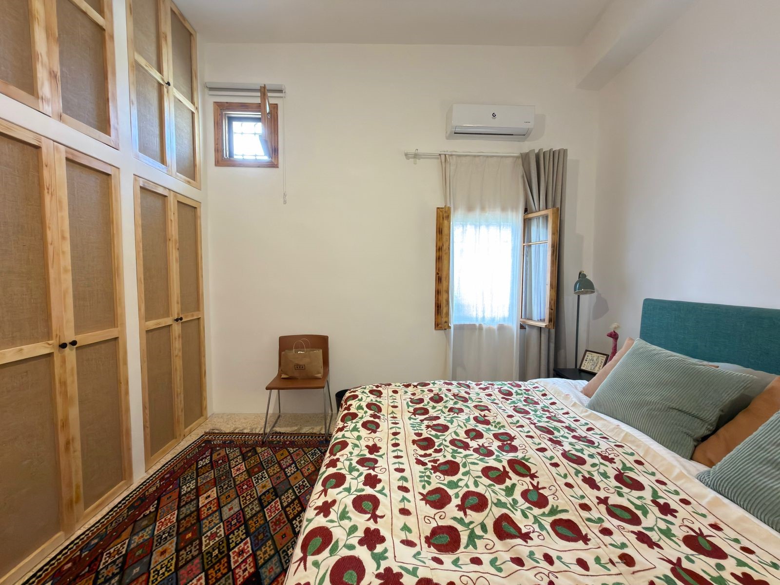 bf29e28b-ff12-4d29-9169-1f84cf767a3e Bedroom with a large wooden wardrobe along the left wall, a bed with a red/green floral quilt, and a small chair nearby.