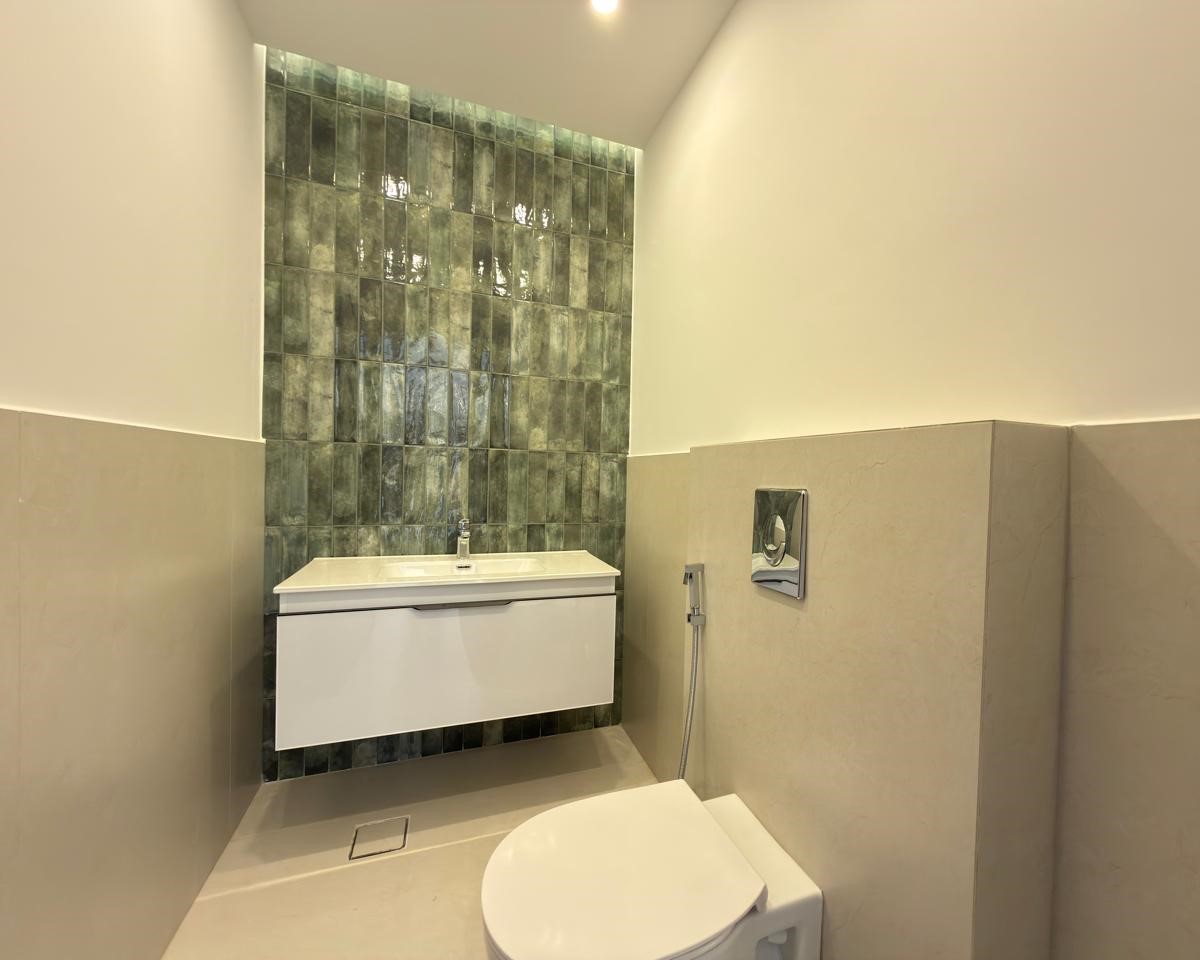 c12c4a7e-7a21-40bc-9f4a-0fc7f8c26cf2 Modern bathroom with a green tiled accent wall, white wall-mounted vanity, and a toilet with a chrome flush plate and handheld bidet spray in a beige-tinted room.