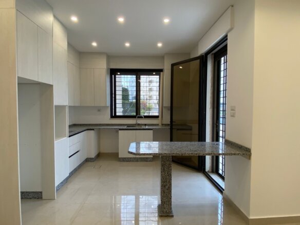 Modern white kitchen with granite countertops, tall cabinets, and an island; large window and sliding glass door to the outside world of the home interior.