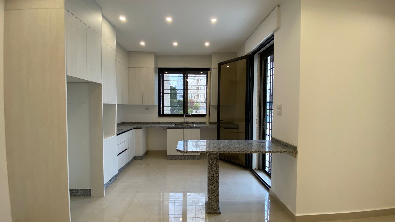 c3672b81-6239-495d-a83a-7f0c016e78b2 Modern white kitchen with granite countertops, tall cabinets, and an island; large window and sliding glass door to the outside world of the home interior.