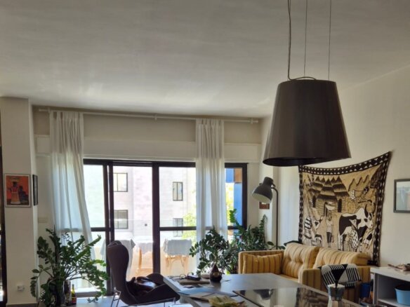 Open-plan living area with a large pendant lamp over a kitchen island, a striped yellow sofa, tapestry wall art, and potted plants by the glass balcony doors.