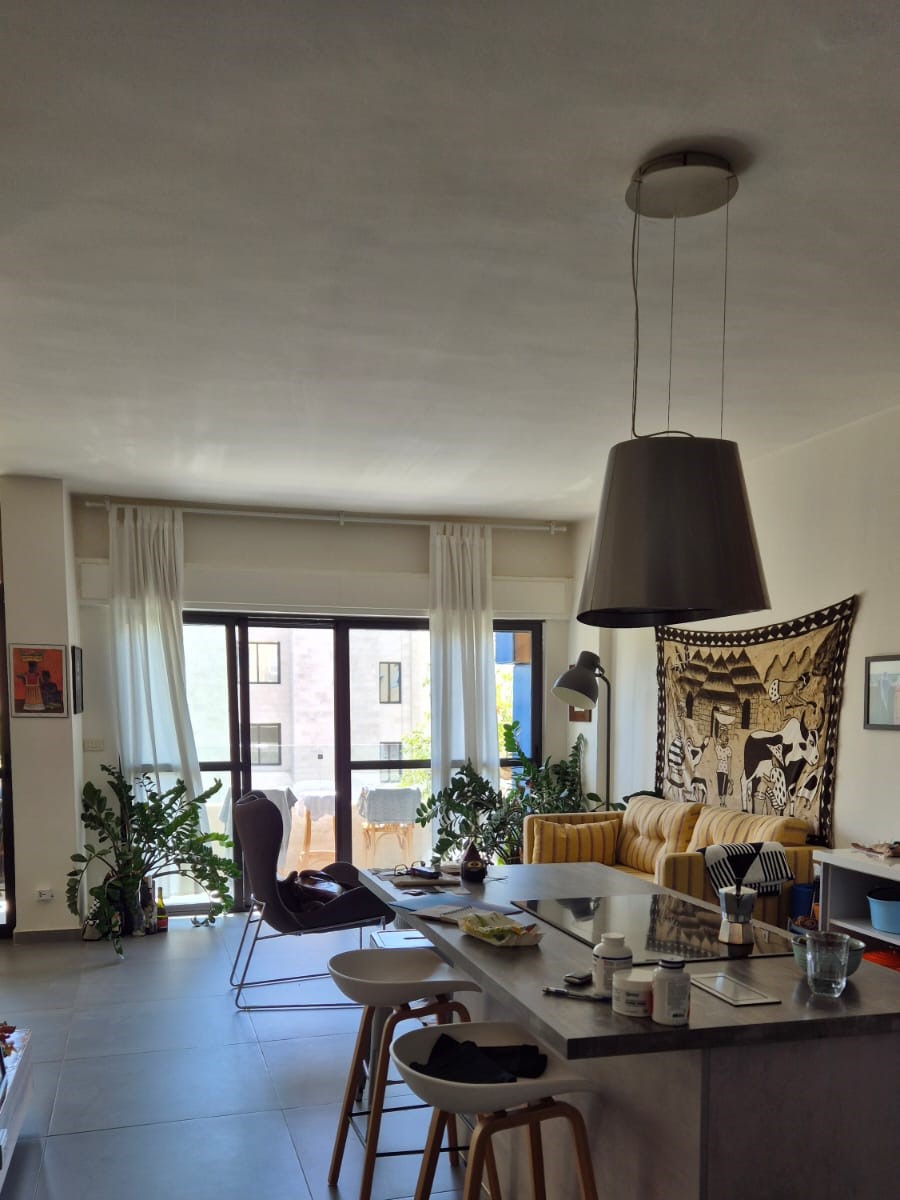 Open-plan living area with a large pendant lamp over a kitchen island, a striped yellow sofa, tapestry wall art, and potted plants by the glass balcony doors.