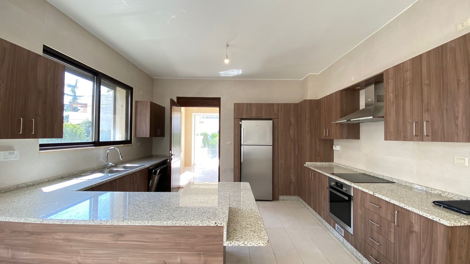 c47b8df0-e0c0-471a-bbbc-3fd1885ee9c2 Modern L-shaped kitchen with wood veneer cabinets, granite countertops, and stainless steel appliances including fridge, oven, and range hood; large window over the sink.